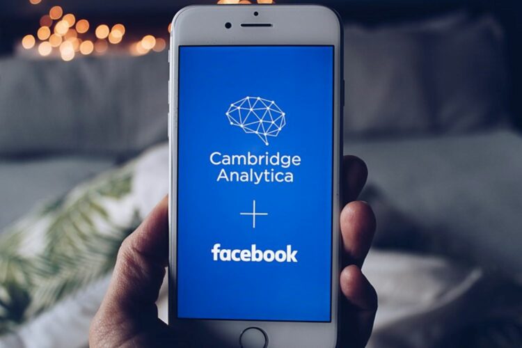 Pantalla de móvil con el logo de Cambridge Analytica y el nombre de Facebook sobre fondo azul cobalto