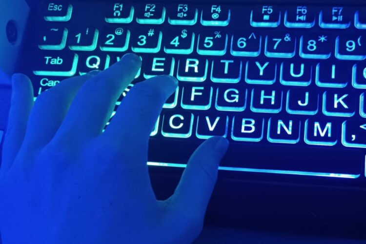 Una mano izquierda iluminada en luz azul cobalto toca un teclado negro con letras iluminadas en blanco.