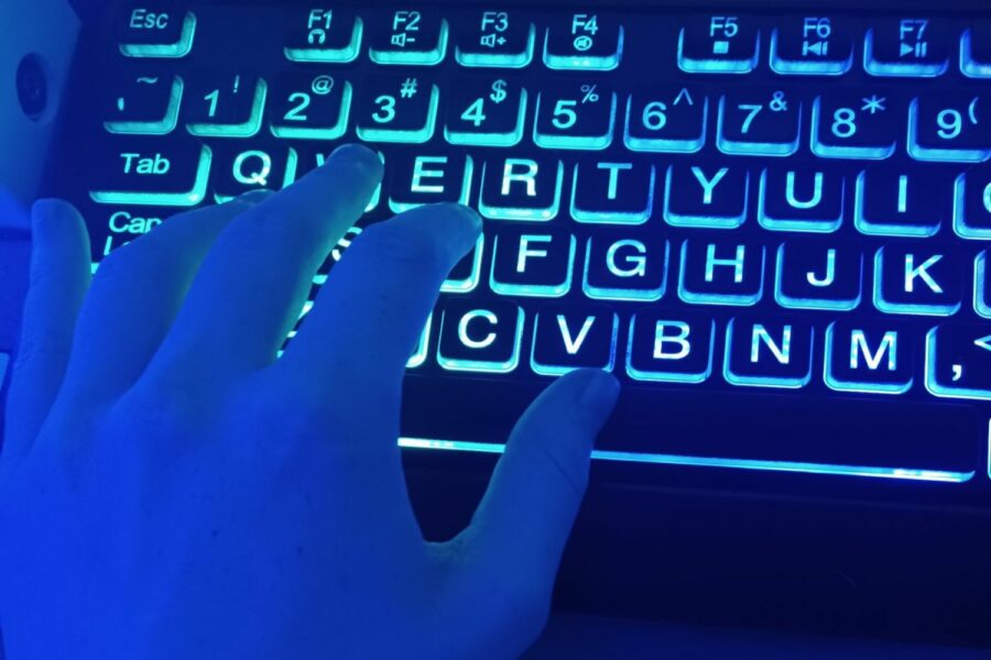Una mano izquierda iluminada en luz azul cobalto toca un teclado negro con letras iluminadas en blanco.