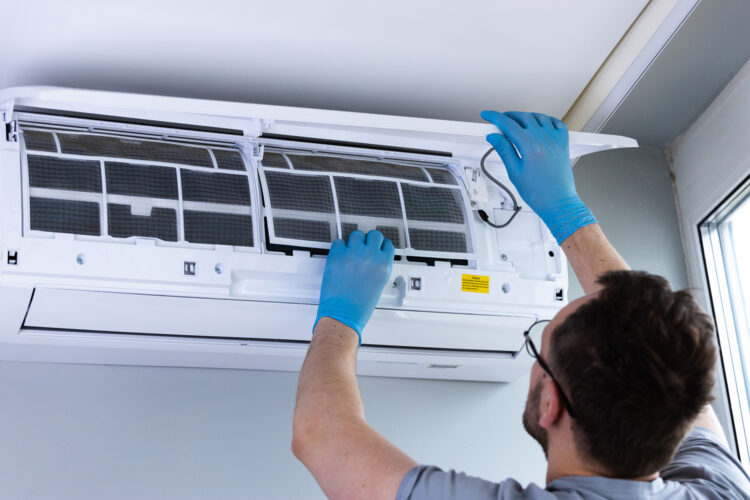 An HVAC technician works on an air conditioning unit installed on the wall, focusing on cleaning or replacing the filter to improve air quality and system performance indoors.